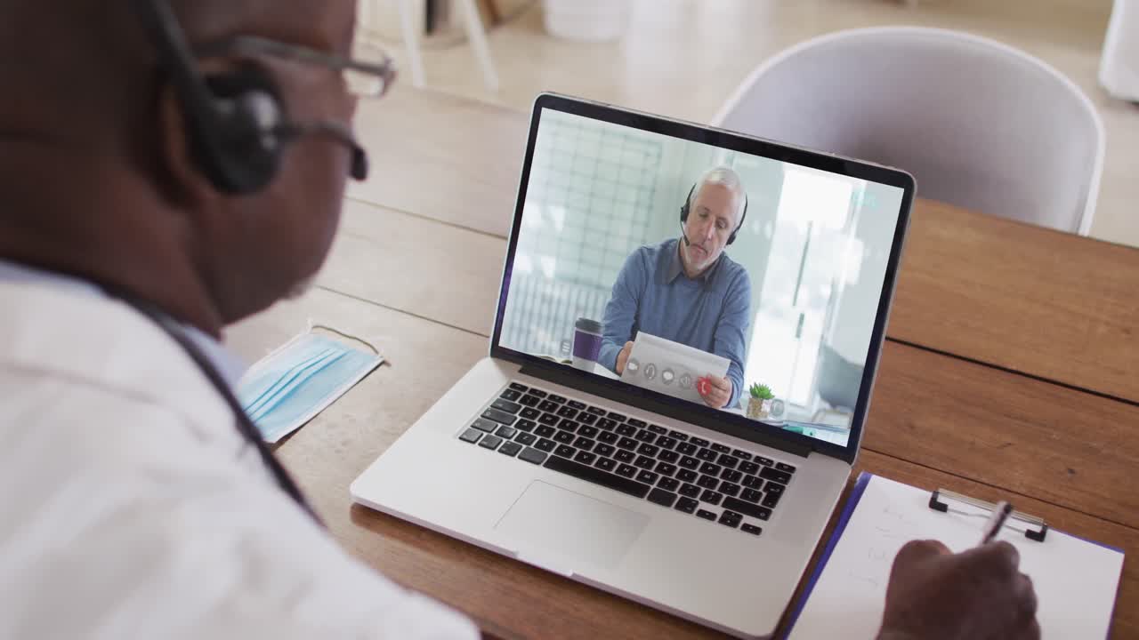 Ein afroamerikanischer Arzt mit Telefon-Headset macht sich Notizen während eines Videoanrufs am Laptop.
