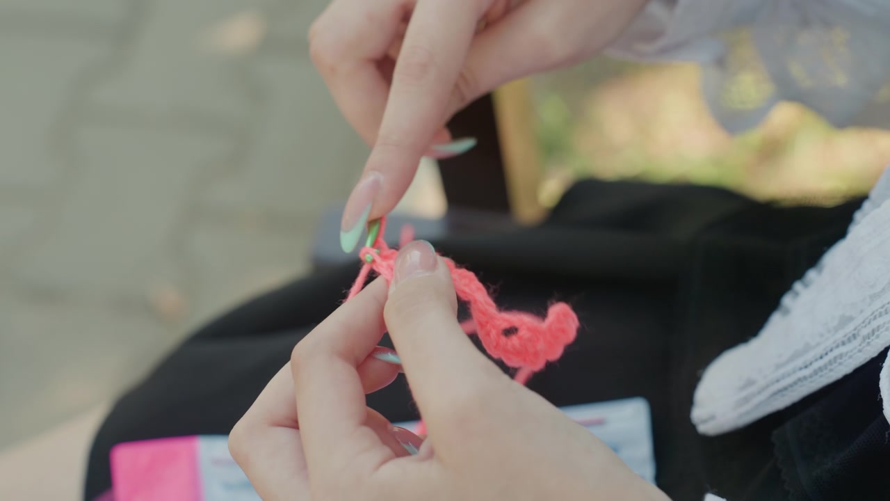 Primo piano di mani che lavorano all'uncinetto con filato rosa e uncinetto verde, mostrando un lavoro tessile dettagliato. La persona manipola continuamente il progetto all'uncinetto in un ambiente esterno con luce naturale soffusa.