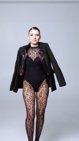 stylish blonde female wearing black bodysuit and lace outfit walks in studio. model puts on a black jacket. vertical video.