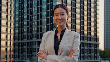 young asian businesswoman wearing professional suit standing confidently with crossed arms, smiling in front of modern office buildings within bustling metropolitan cityscape