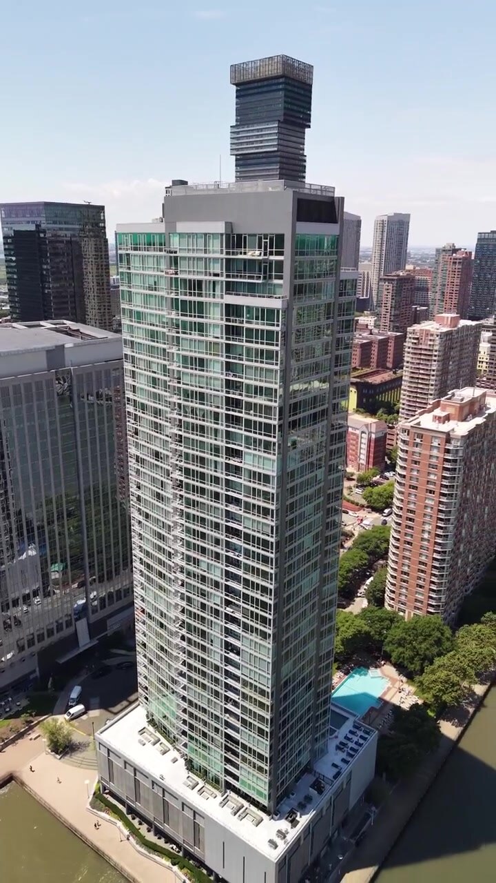 Vista aérea de um arranha-céu residencial contemporâneo na orla do rio Hudson em Jersey City, com o horizonte do centro da cidade e a arquitetura urbana circundante visíveis durante um dia ensolarado.