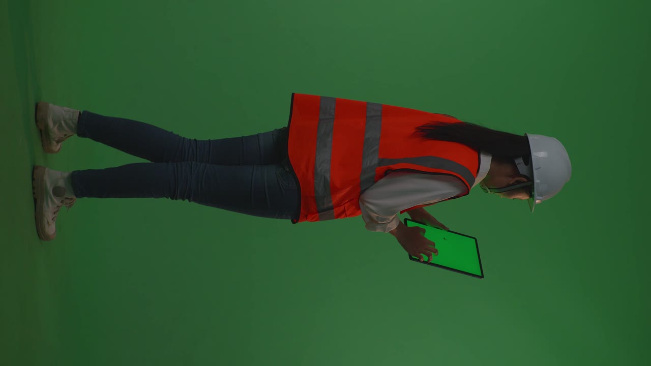 vista trasera de todo el cuerpo ingeniera femenina asiática con casco de seguridad trabajando en una tableta de pantalla verde mientras está de pie en el estudio de fondo de pantalla verde, durante la puesta o salida del sol