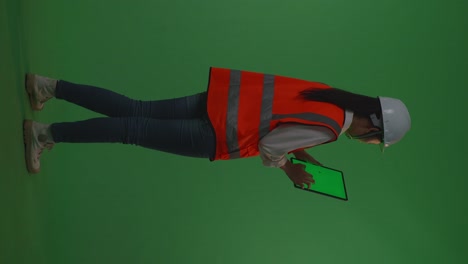 back view of full body asian female engineer with safety helmet working on a green screen tablet while standing in the green screen background studio, during sunset or sunrise time