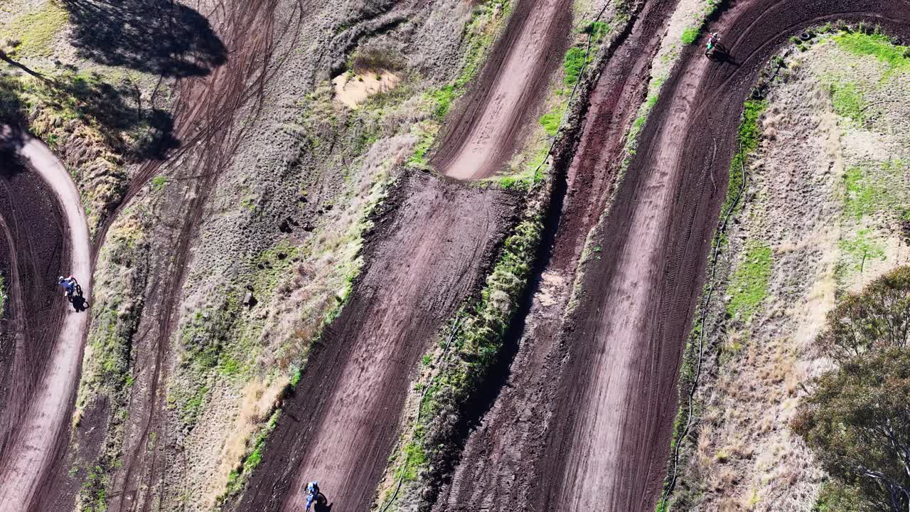 Motorbikes speed around a winding dirt track, captured from above in bright daylight