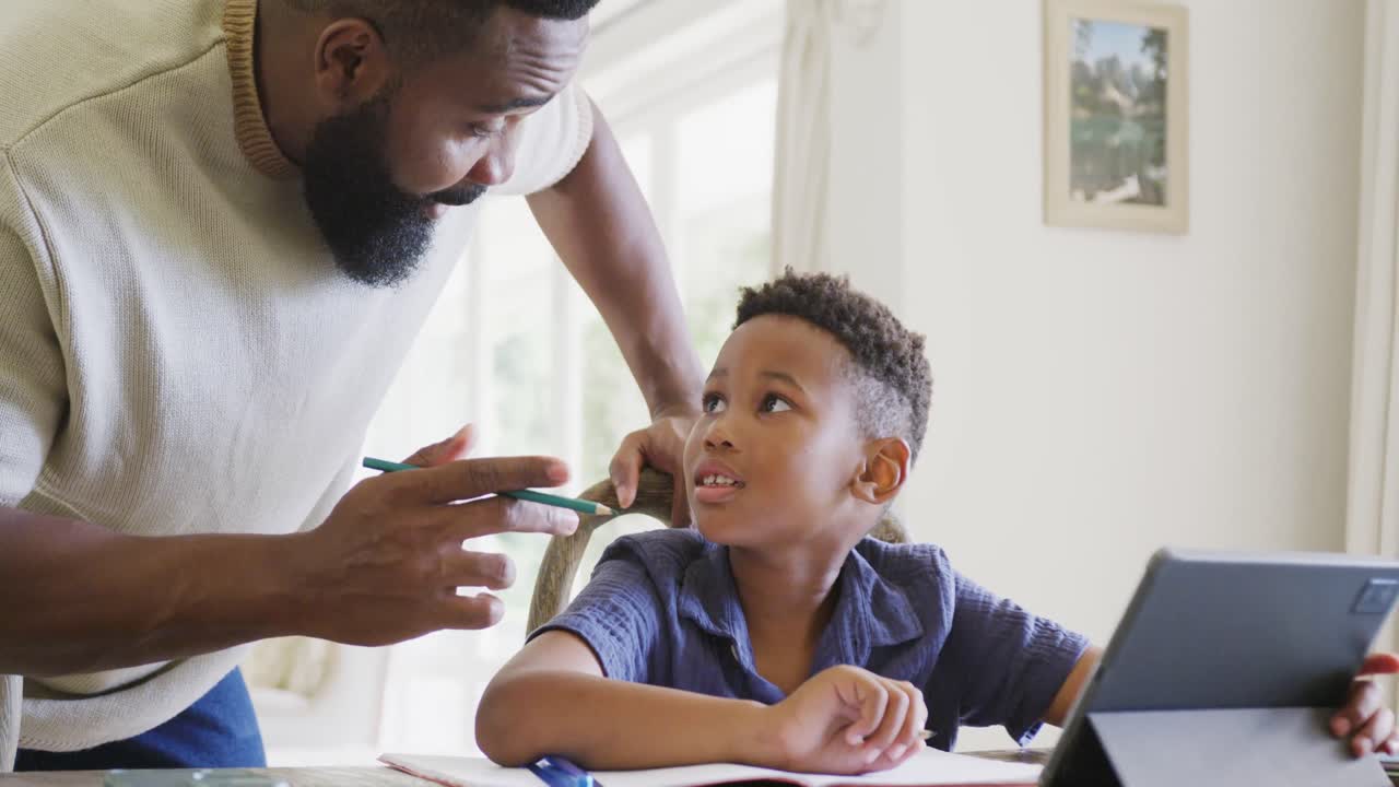 Happy african american father and son sitting at table and doing homework together, in slow motion