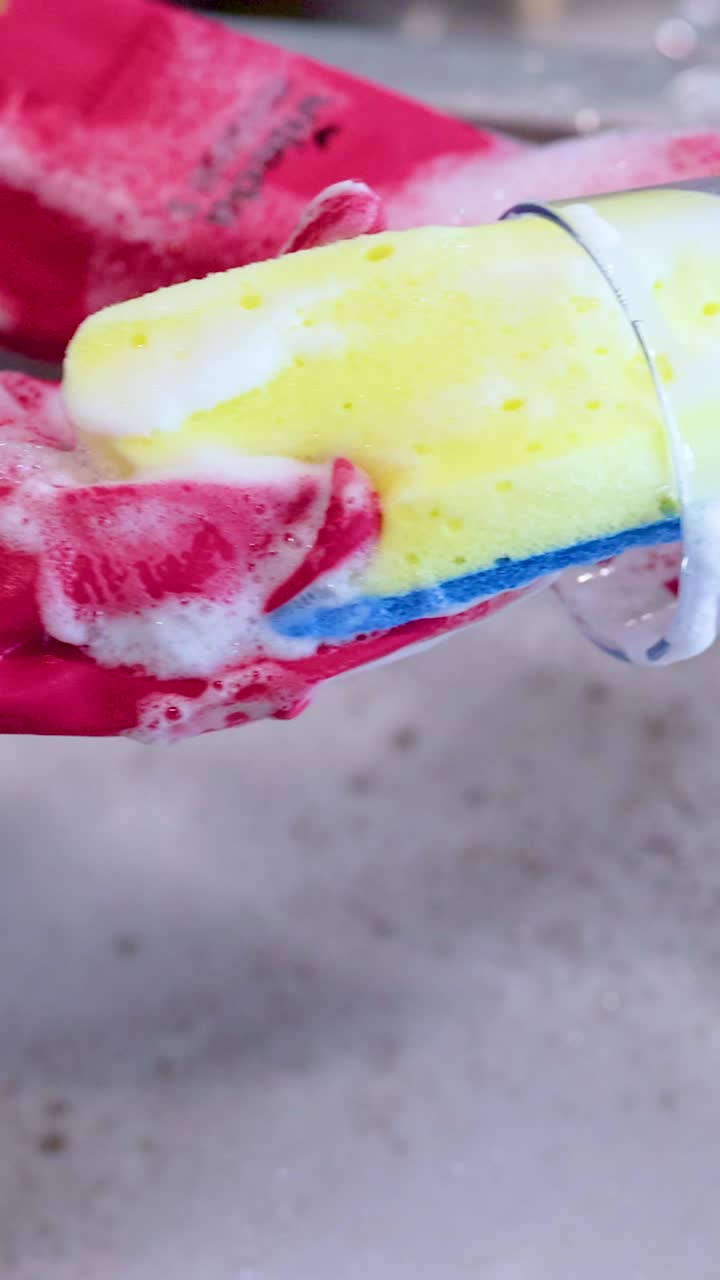 Hands in red gloves clean a glass with a soapy sponge in a kitchen sink