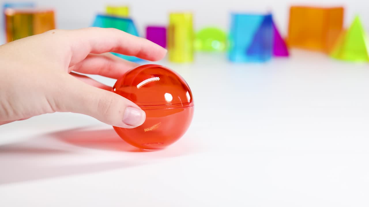 A hand interacts with a red sphere on a table surrounded by colorful geometric shapes in bright lighting