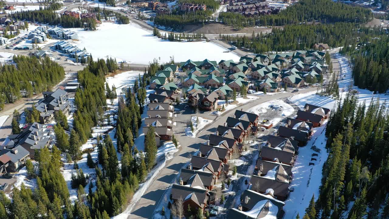 Bird’s eye drone view of Big Sky, Montana shows clusters of vacation condos and snowy streets set among forested hills and framed by dramatic peaks in a peaceful alpine landscape