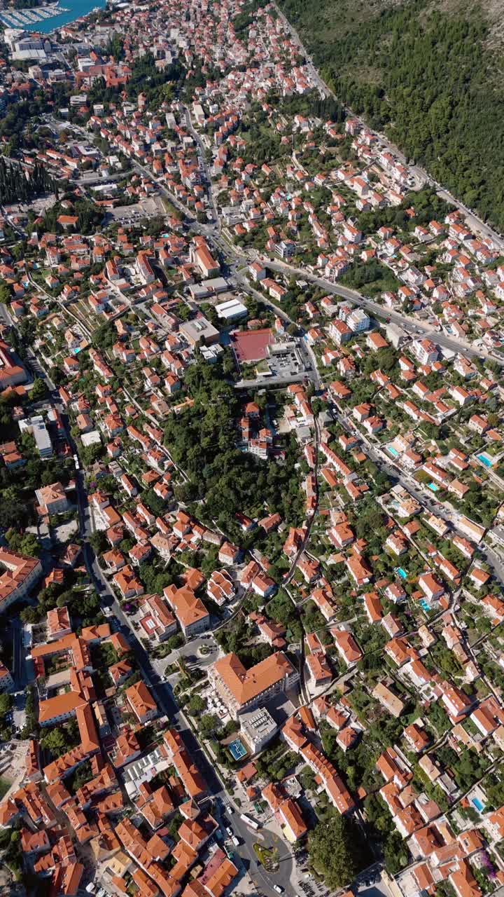 Aerial Birds Eye drone shot over Dubrovnik, capturing red rooftops, scattered trees, the coastal port with docked boats, and light traffic winding through streets along the historic cityscape