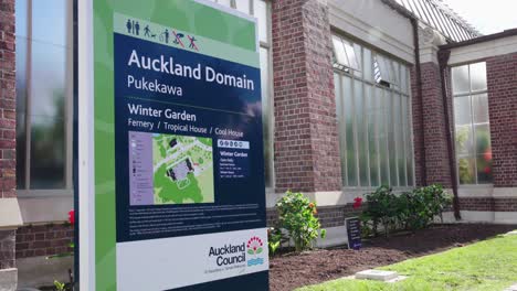 still view of an auckland domain sign with information about the winter garden fernery, tropical house, and cool house in front of a brick and glass building