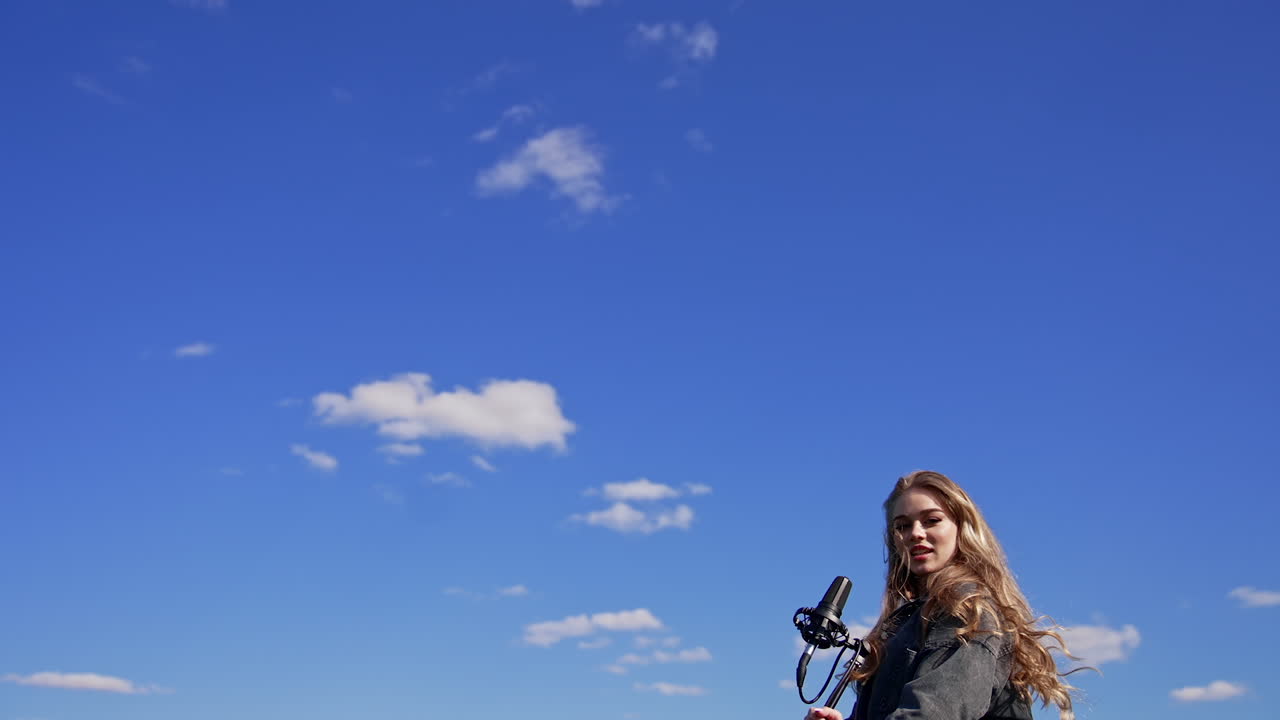 Beautiful female performing a song alone outdoors. Young singer girl with a microphone on blue sky background. View from below.