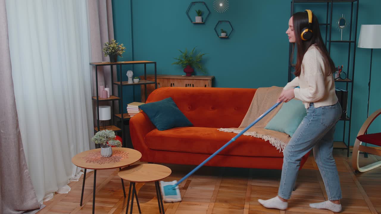 Joyful young woman is mopping floor in apartment living room, having fun listening to music at home