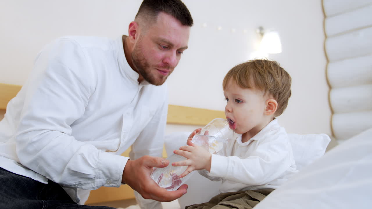 Caring father takes a water bottle to his baby boy. Little kid sitting on the bed tries to drink water himself.