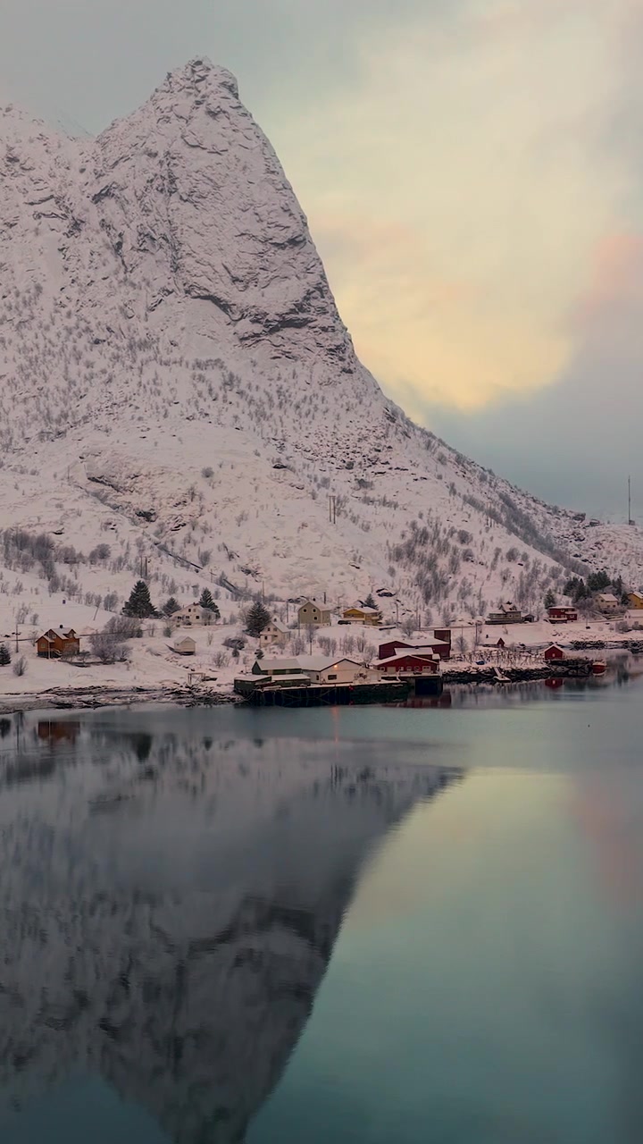 Et fantastisk filmatisk luftfoto av den snødekte fiskerlandsbyen Reine i Lofoten, Norge, med rolige vannrefleksjoner og majestetiske fjell under den gylne timen.