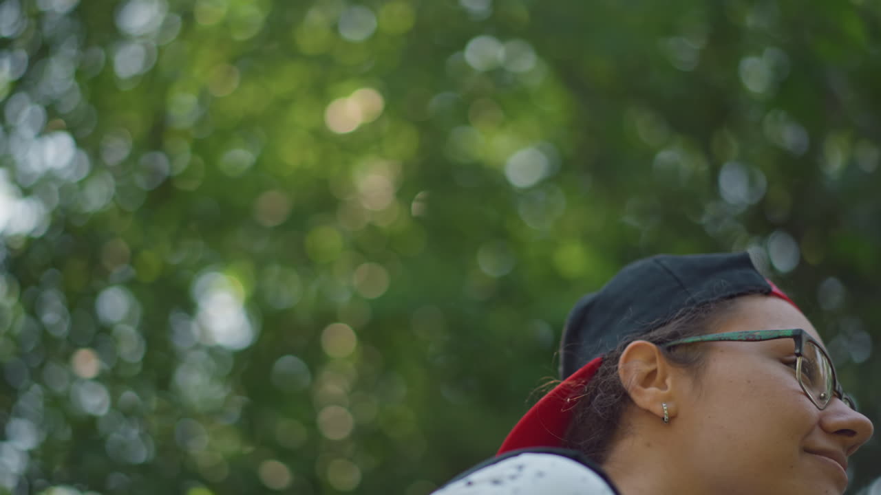 나무 아래에서 배낭을 멘 젊은 사람이 쾌활한 트레커 분위기와 브이로그를 찍는 듯한 에너지로 햇빛이 비치는 캐노피를 올려다보고 있어요. 솔직한 미소와 활기찬 걸음, 배낭끈과 캐주얼한 옷차림, 밝은 녹색이 돋보입니다.