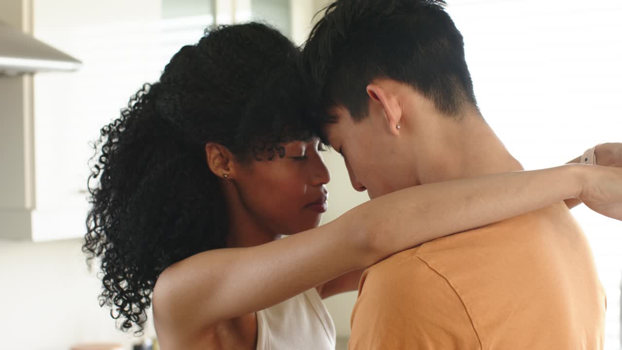 Couple in home kitchen, African American woman initiating embrace, soothing Asian man by window
