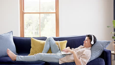 happy biracial man wearing headphones lying on couch using laptop at home, copy space, slow motion