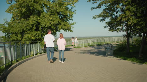 lady in pink top and jeans walking alongside her friend holding laptop in left hand near iron rail on sunny paved pathway with greenery and benches, two women walking ahead, framed by trees