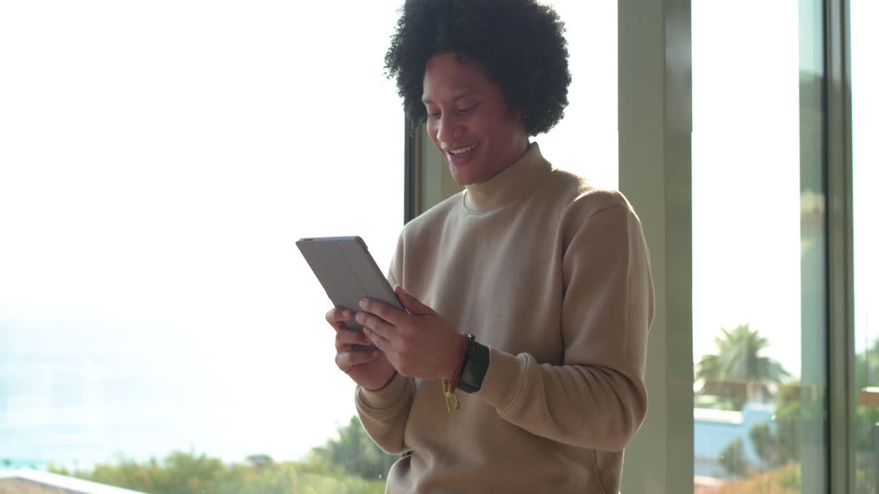 Mid adult male reading tablet and swiping for work while leaning toward coastal balcony windows