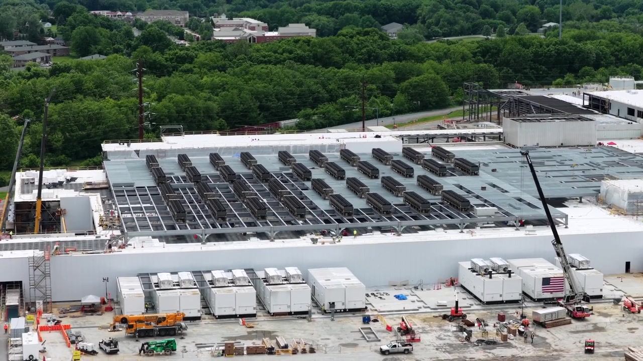 Enorme cantiere di un data center con unità di raffreddamento sul tetto e gru, bandiera americana vicino ai generatori in un ambiente boschivo. Lancaster, Pennsylvania, USA. Ampia inquadratura aerea con drone.