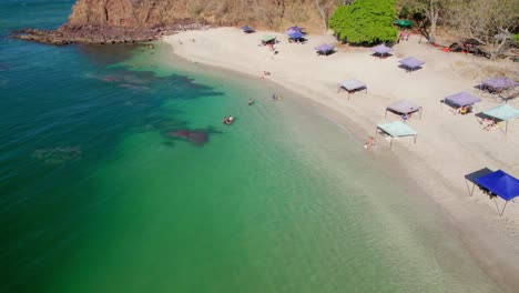 Popular-Busy-Beach-Shore-With-Tourists-Relaxing-In-Shade-And-Swimming,-Conchal-Beach-Costa-Rica-4K-Drone