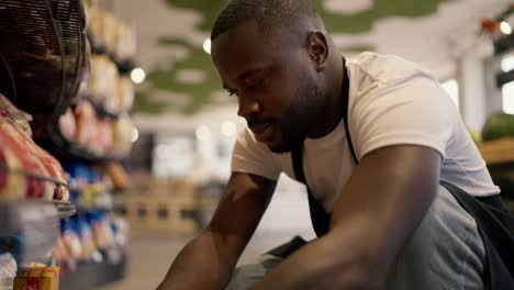 close-up shot of a black-skinned man in a white t-shirt and black apron crouching near a shop window and laying out goods in a large supermarket