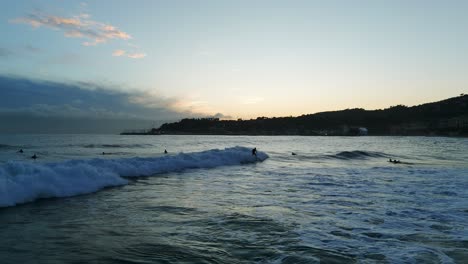 Aerial-view-of-surfers-riding-waves-at-sunset-along-Varazze-coastline-in-Luguria,-Italy