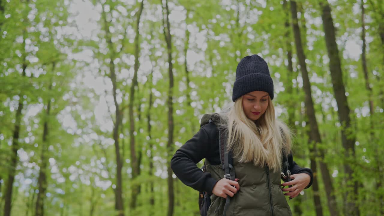 A young woman wearing a beanie and backpack looks up at the tall trees while hiking through a lush green forest. The scene captures a moment of curiosity and connection with nature during an adventure