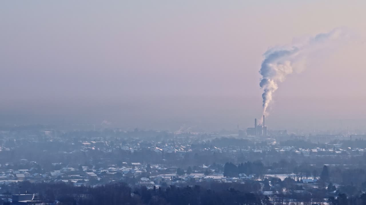 Una colonna di fumo si alza da un impianto industriale sopra un paesaggio urbano nebbioso sotto un cielo invernale pallido - Ripresa aerea con drone