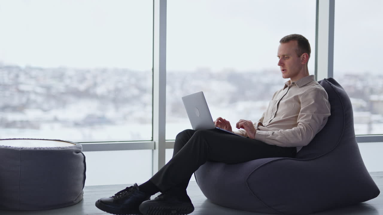 Busy young adult man works on laptop sitting in a chair. Comfortable working atmosphere in spacious room with panoramic windows.