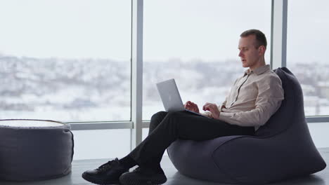 busy young adult man works on laptop sitting in a chair. comfortable working atmosphere in spacious room with panoramic windows.