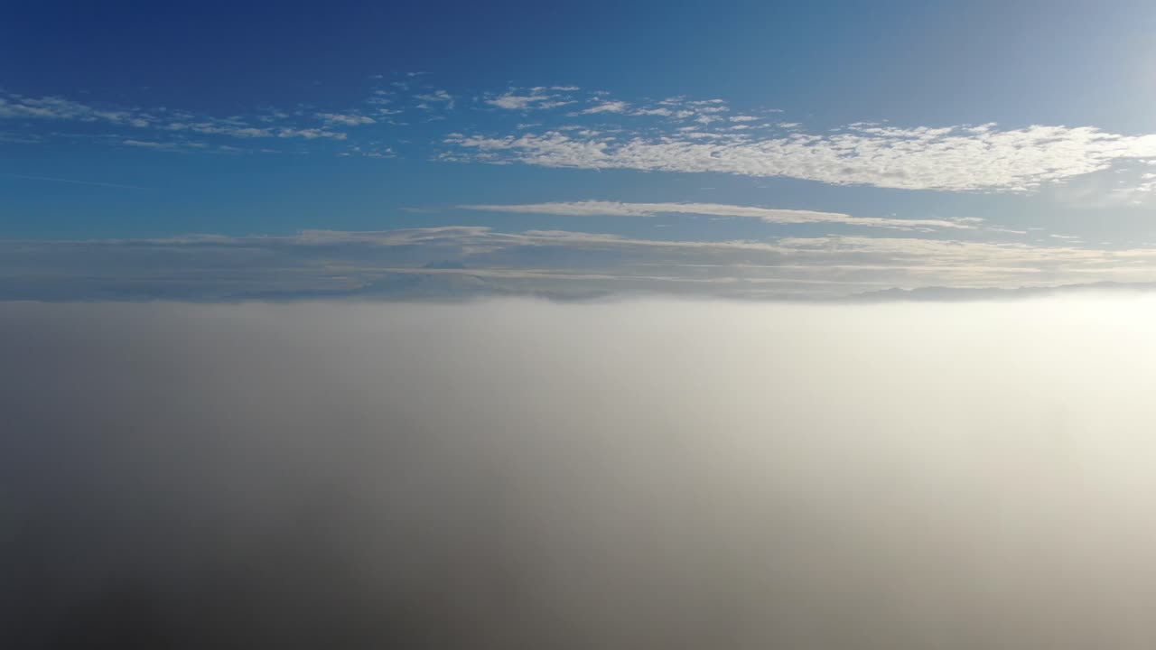 Drone rises over a foggy landscape on a beautiful blue sky day with mottled clouds to reveal a mountain vista