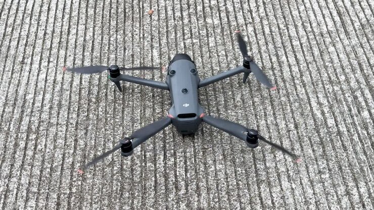 Um plano em ângulo alto captura um drone quadricóptero cinza acionando suas hélices em concreto texturizado.