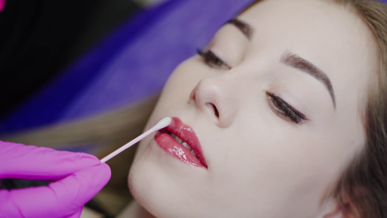 Permanent lips make-up. Cosmetologist finishes the cosmetic procedure on lips to a lovely female client. Close-up.