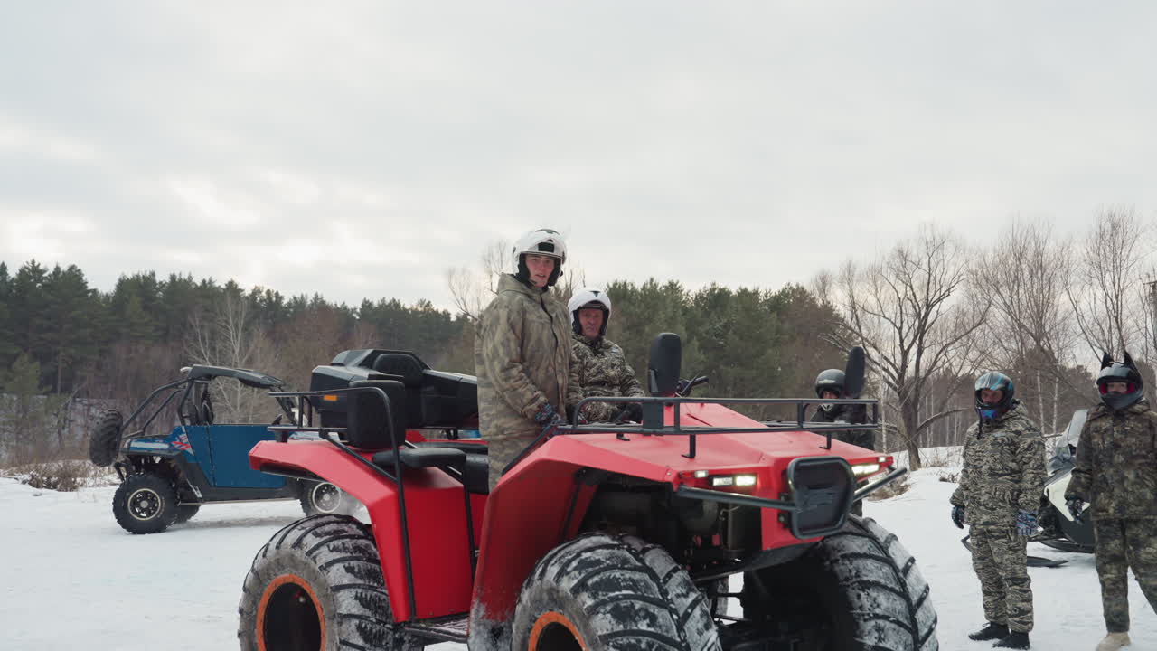 Primo piano di un quad rosso con piloti che preparano l'attrezzatura e controllano i mezzi, convoglio di quad e motoslitte dietro, il leader che sistema il casco e la mappa, campo invernale nuvoloso, atmosfera concentrata prima dell'azione estrema