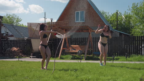 two women in black bikinis playfully chase each other across green lawn as one sprays water from hose, droplets sparkling in warm sunlight, laughter echoing under blue sky
