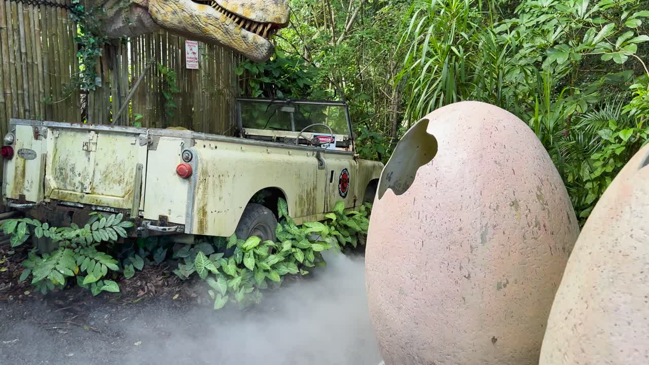 Animatronic dinosaur hatches from egg near vintage vehicle, surrounded by dense greenery and mist