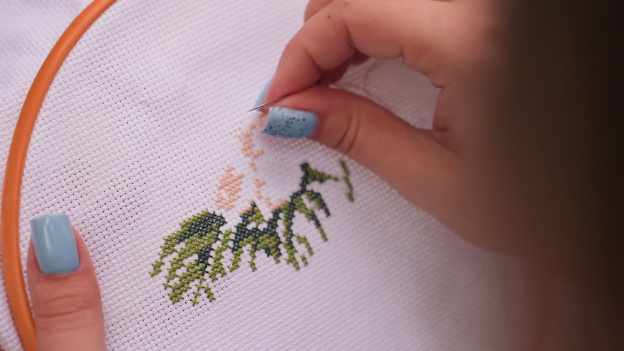Close up hand view of woman with painted blue nails working on detailed embroidery pattern using thread and needle, stitching into fabric hoop under natural light with calm focus