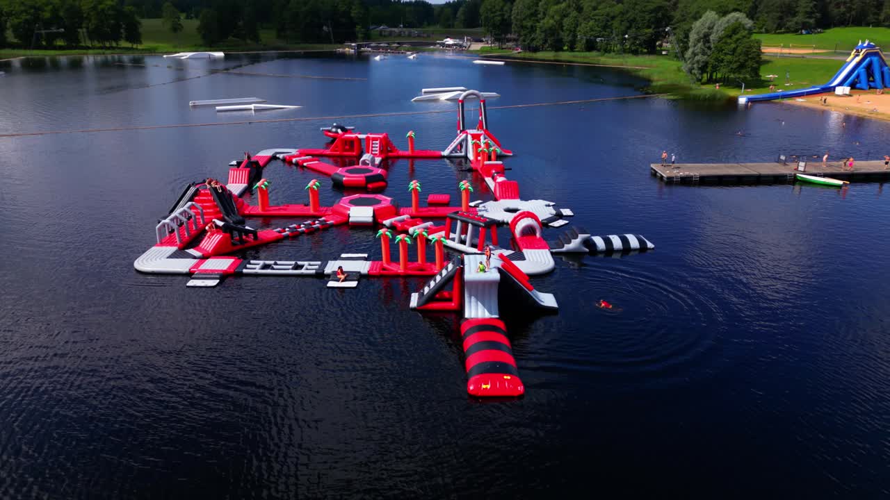 Colorful floating water park with red inflatables on Zarasai lake, with swimmers, pier, and forest backdrop. Shot in Zarasai, Lithuania (Zarasai, Lietuva)
