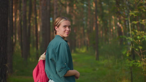 mujer de pie en el bosque, juguetón besando el aire con una sonrisa, con camisa verde y llevando una mochila roja, ella irradia alegría despreocupada rodeada de árboles