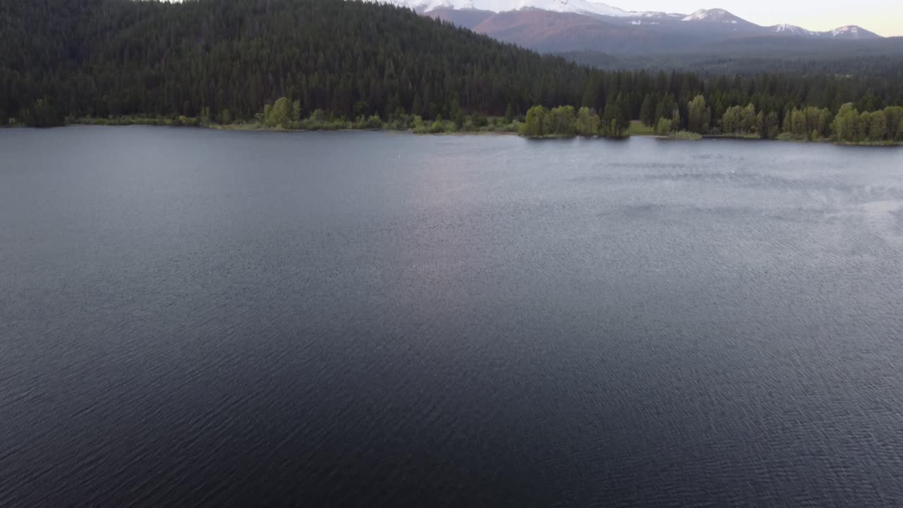 Reveal of Mount Shasta volcano in California from the water of Lake Siskiyou at sunset. Reveals white snow-capped peak of the mountain with clear skies and sunset colors. Filmed with DJI drone in 4K.