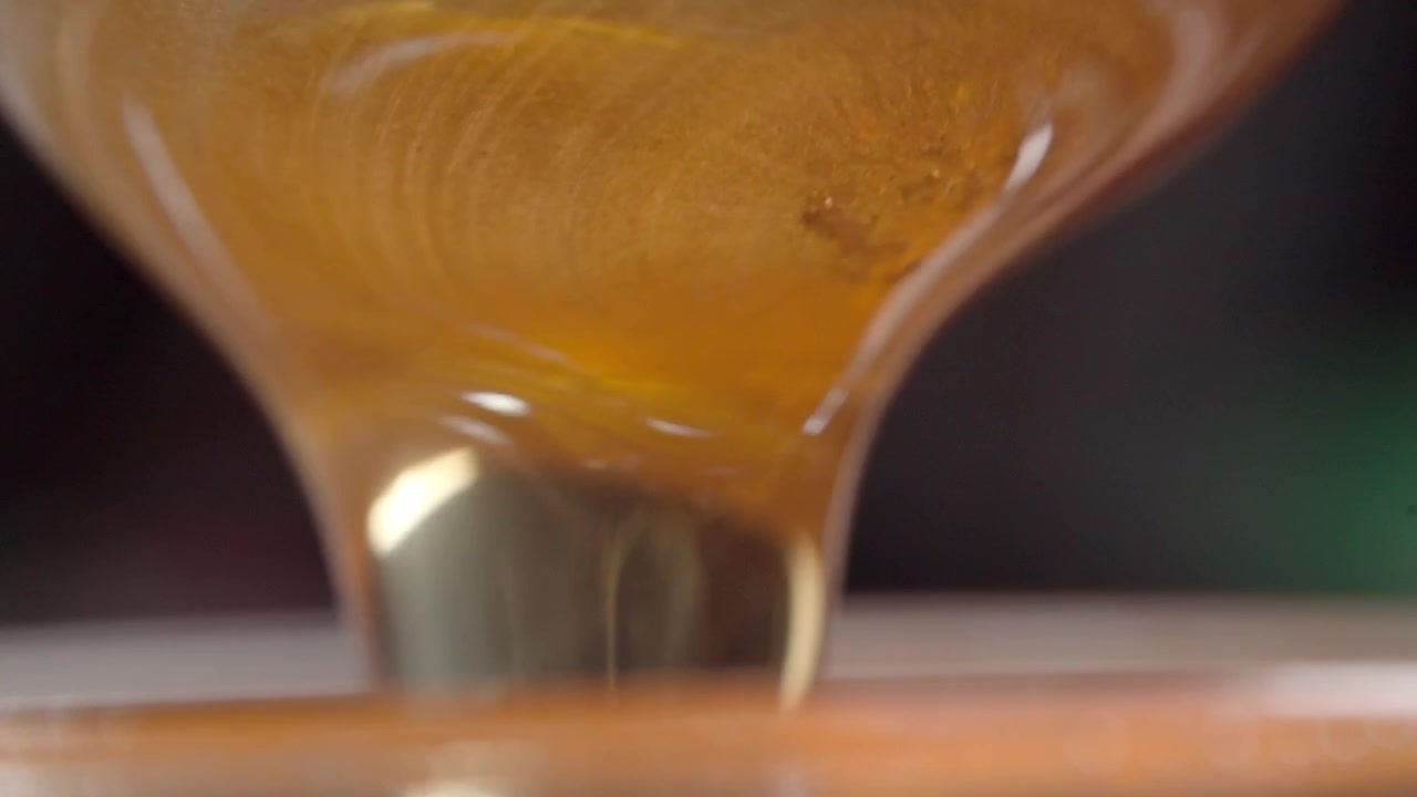 Primo piano macro di miele denso che scorre su un mestolo di legno per miele al rallentatore. Il miele dorato ricopre la superficie scanalata, evidenziando la consistenza liscia, la viscosità e la dolcezza naturale.