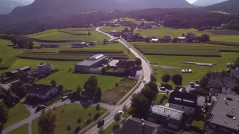 Drone-flying-at-higher-altitude-into-the-sun-over-streets-cars-and-houses-and-companys-towards-fields-of-green-and-mountains-in-the-back-of-the-Alps