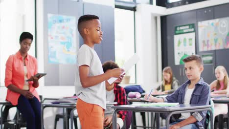 Diverse-female-teacher-and-happy-schoolchildren-at-desks-reciting-in-school-classroom