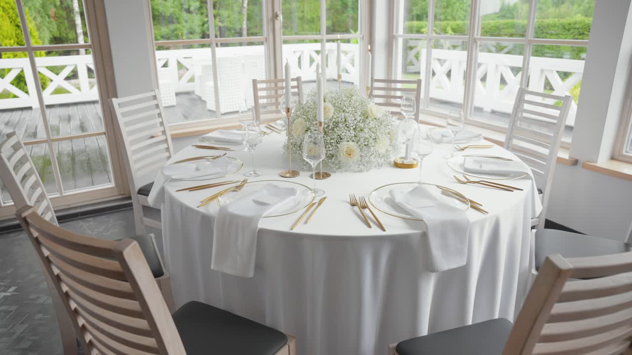 Elegant indoor wedding reception hall with white-decorated tables, floral centerpieces, and large windows overlooking lush greenery, creating a bright and romantic atmosphere