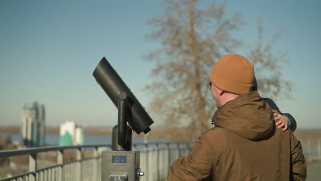 a father holds his son close near a public telescope, playing together with a cityscape in view, the father, wearing a brown jacket and his son wears a black jacket