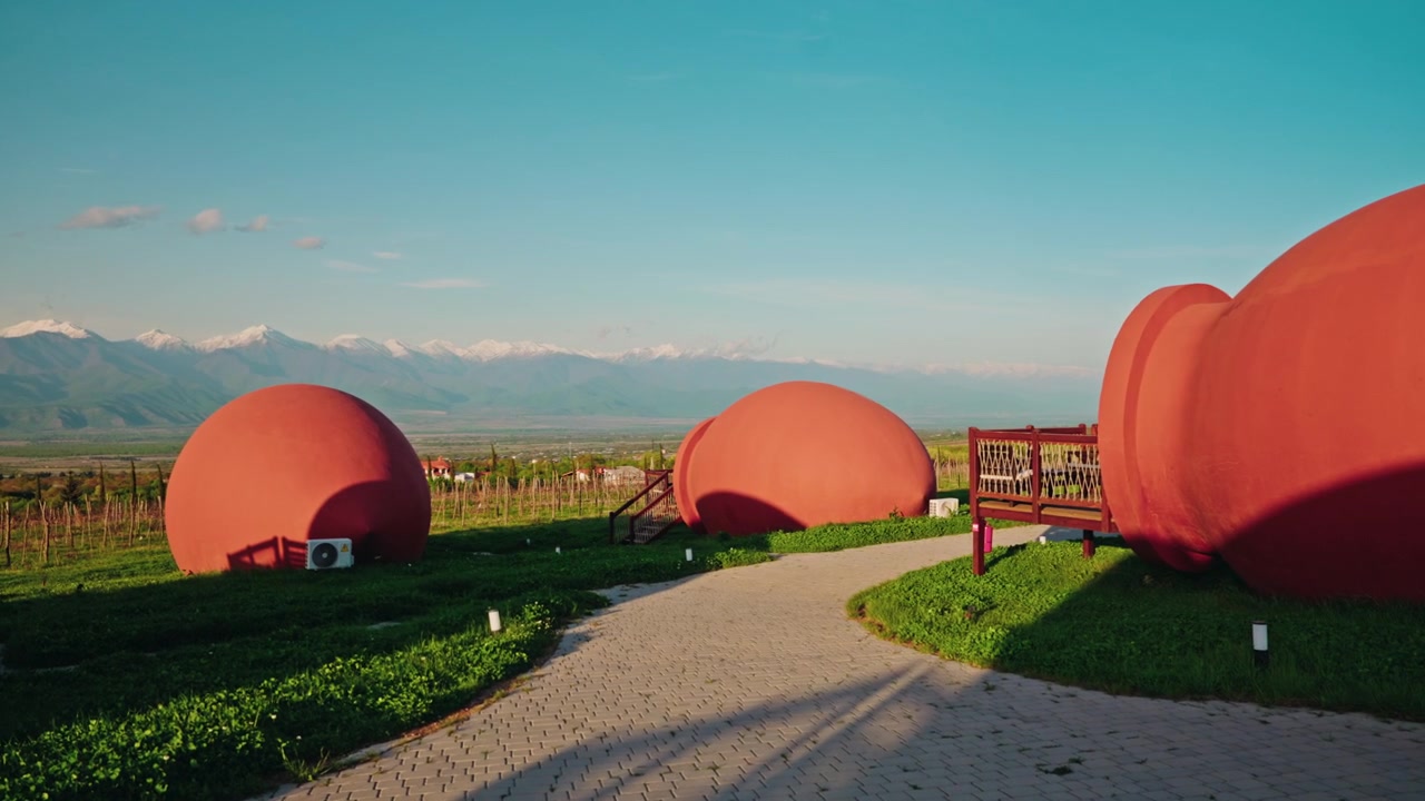 File di camere d'albergo a forma di qvevri in terracotta si trovano su una collina verde sopra un vigneto, con il Caucaso innevato all'orizzonte
