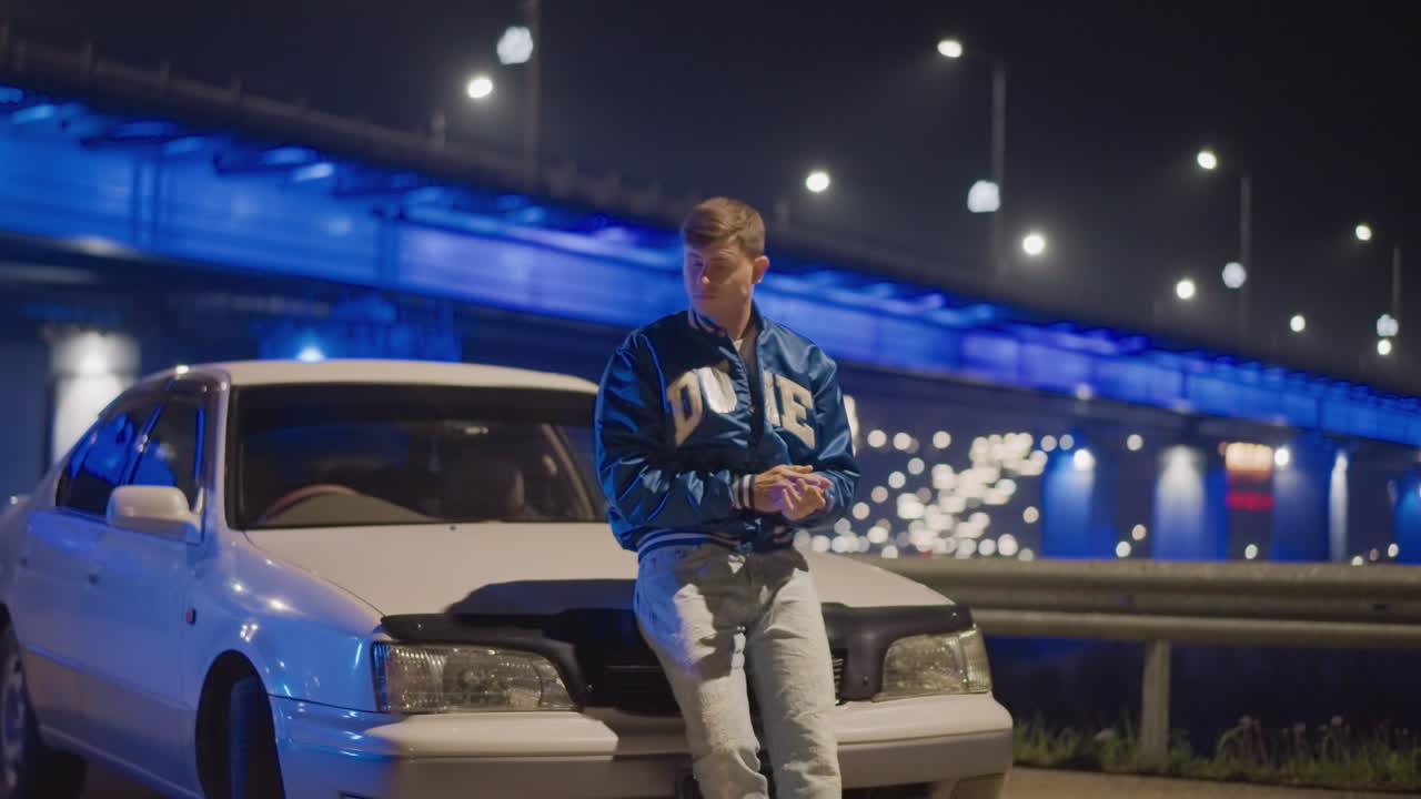 Puente nocturno con coche y un hombre blanco apoyado en el capó bajo luces de neón azules, bokeh cinematográfico, chaqueta universitaria, berlina aparcada junto a la barrera de protección, conductor nocturno mirando el teléfono, mirador junto a la carretera, ambiente reflexivo