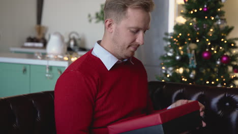 blond man opens a christmas present sitting on the sofa in a living room with christmas decorations