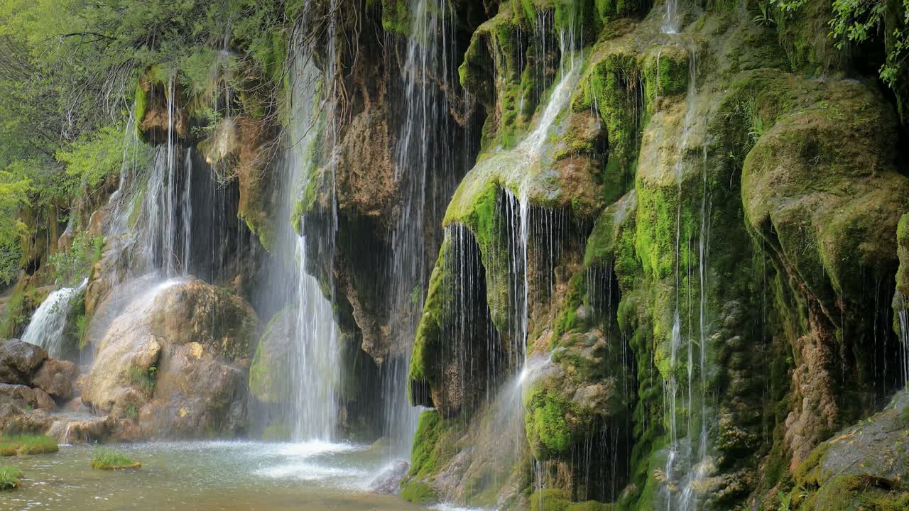 Vista cinematografica di una cascata che scorre delicatamente su una rupe rocciosa ricoperta di muschio con sottili ruscelli e una leggera nebbia che creano un'atmosfera serena, una texture ricca e un pacifico scenario naturale